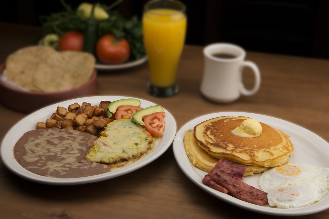 Classic American breakfast of pancakes, bacon, and eggs alongside a tex-mex breakfast of beans, eggs, tomatoes, avocados, and home fries with a class of orange juice and cup of coffee in the background