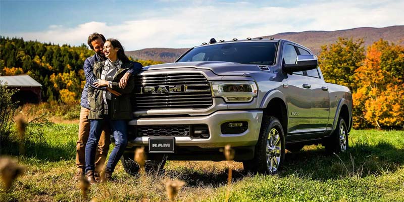 A couple leaning against the grill of a silver 2024 RAM 2500 on a sunny day with beautiful orange and yellow trees in the distance